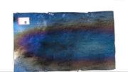 A rectangular metal panel with a dark blue, purple, and bronze heat-tinted finish, leaning against a rusty metal structure outdoors.
