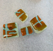 Several small rectangular fused glass beads with orange-brown, black, and pale aqua swirls are scattered on a light gray textured surface.