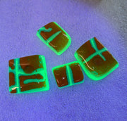 UV-reactive acrylic nail art pieces with a brown base and glowing neon green lines, displayed on a purple-reactive surface with more pieces in the background.