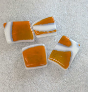 Four square glass tiles with a glossy orange and white marbled pattern, arranged on a textured light gray surface.